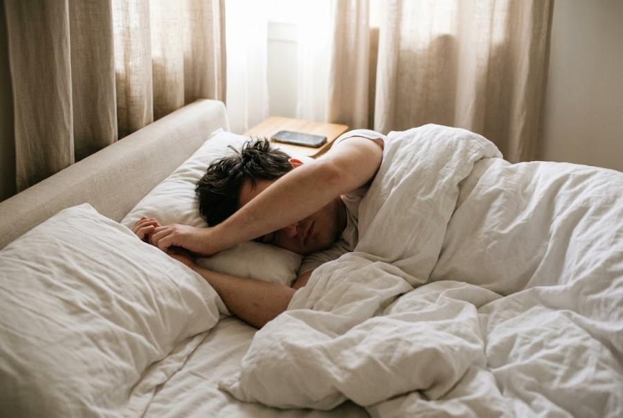 A softly lit bedroom in early morning, curtains half-drawn against the window light, an unmade bed and a phone on the nightstand